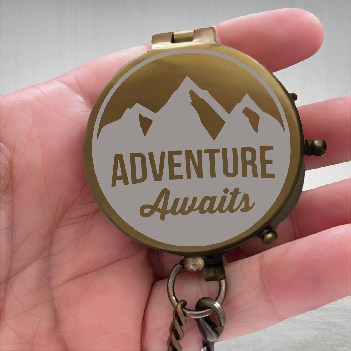 May include: A brass compass with a white background and a mountain silhouette. The compass reads "Adventure Awaits".