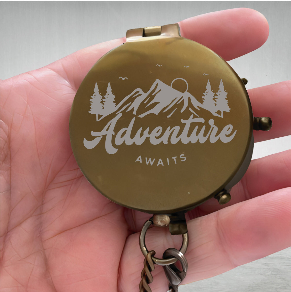 May include: A round, brass-colored metal compass with a hinged lid. The lid features an engraved graphic of mountains, pine trees, and a sun, with the text "Adventure AWAITS" centered below the landscape. A metal ring and chain are attached to the base.