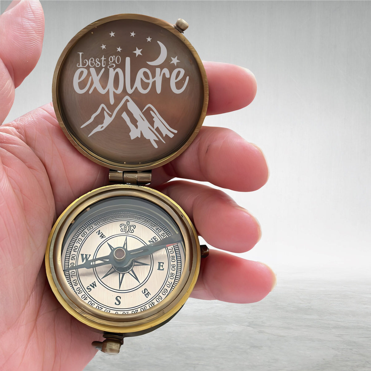 May include: A brass compass with a lid that has the words "Let's go explore" and a mountain range design. The compass face has a black needle and a white background with black markings.