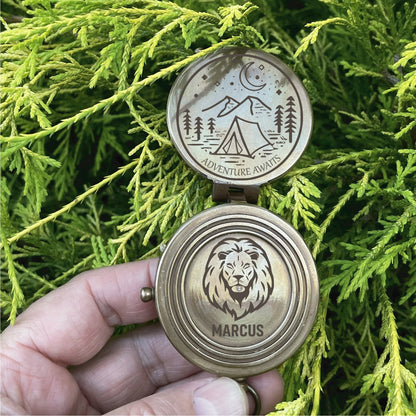 May include: A brass compass with a lion design on the front and a camping scene on the back. The compass reads "Adventure Awaits" and "Marcus".