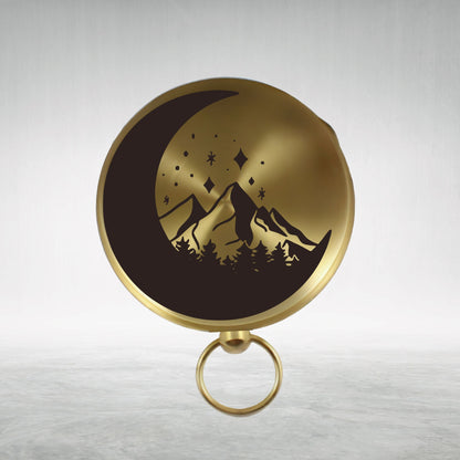 May include: A gold compass with a black design of a crescent moon, mountains, and stars. The compass has a ring handle.