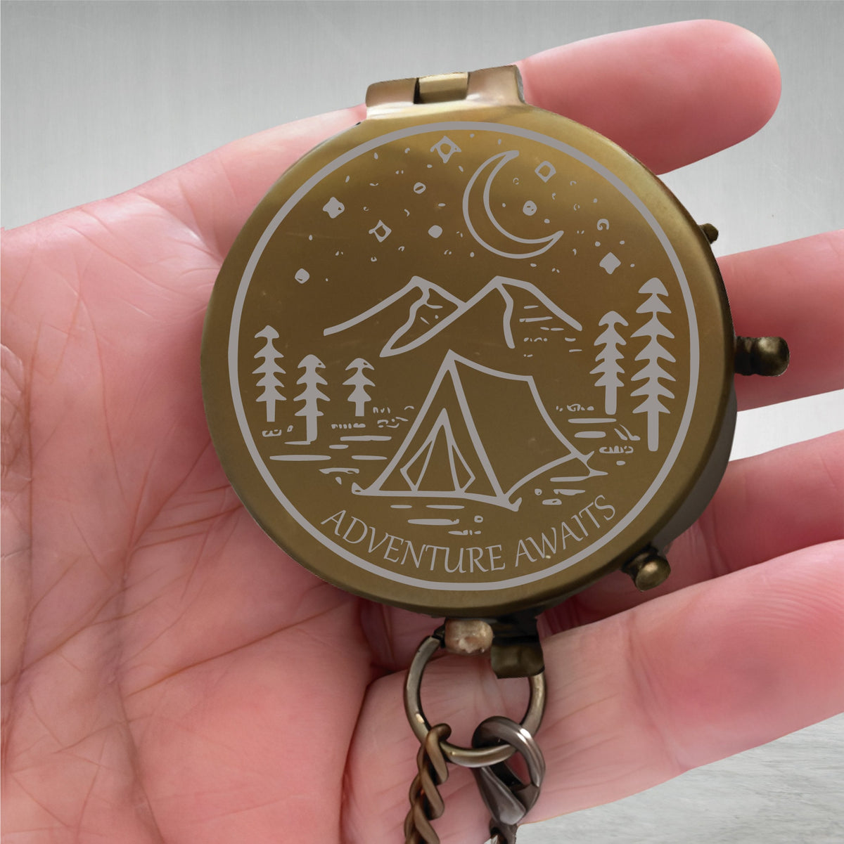 May include: A brass compass with a white engraved design of a mountain range, a crescent moon, stars, and a tent. The compass has a chain attached and the words "Adventure Awaits" are engraved on the bottom.