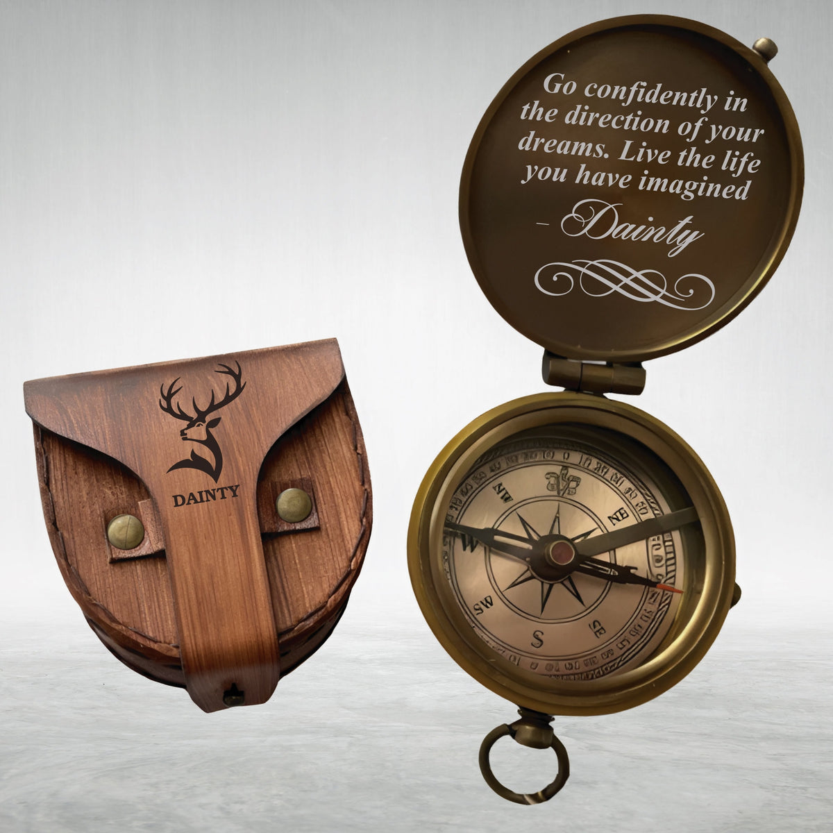 May include: A brass pocket compass with an open lid displaying the text: Go confidently in the direction of your dreams. Live the life you have imagined - Dainty. Beside it is a brown leather pouch featuring a deer head logo and the word Dainty.