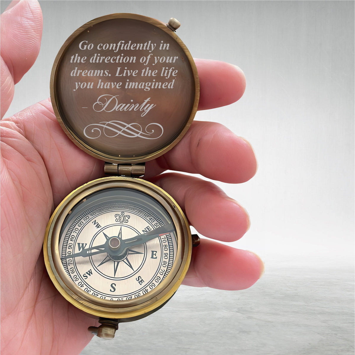 May include: A brass-toned pocket compass with a hinged lid featuring the engraved text: Go confidently in the direction of your dreams. Live the life you have imagined - Dainty. The compass face displays cardinal directions and a central needle.