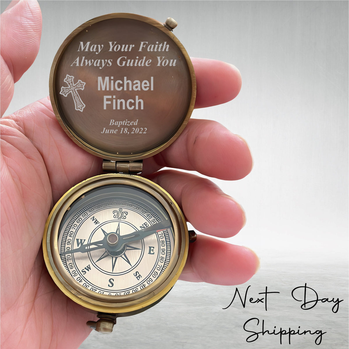 May include: A brass compass with a glass face and a compass needle. The lid of the compass is open and has an engraved message that reads "May Your Faith Always Guide You Michael Finch Baptized June 18, 2022".