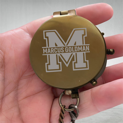 May include: A round, brass-colored pocket compass with a hinged lid. The lid features an engraved large letter M with the name MARCUS GOLDMAN centered horizontally across it. A small metal ring and a portion of a chain are attached to the base.