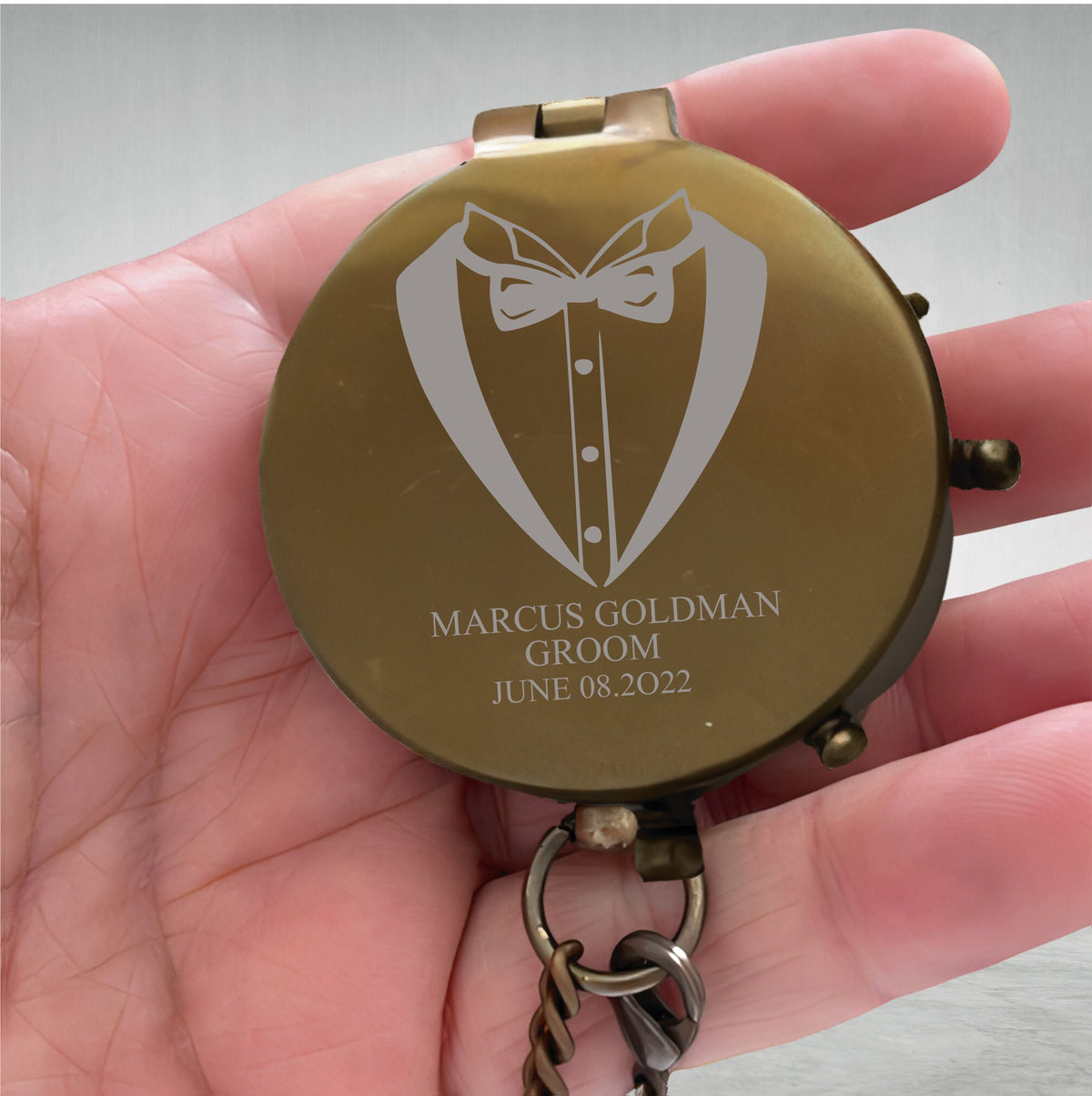 May include: A round, brass-colored pocket compass featuring an engraved tuxedo jacket and bowtie design on the lid. The text MARCUS GOLDMAN GROOM JUNE 08.2022 is etched below the graphic. A metal chain is attached to the base of the compass.