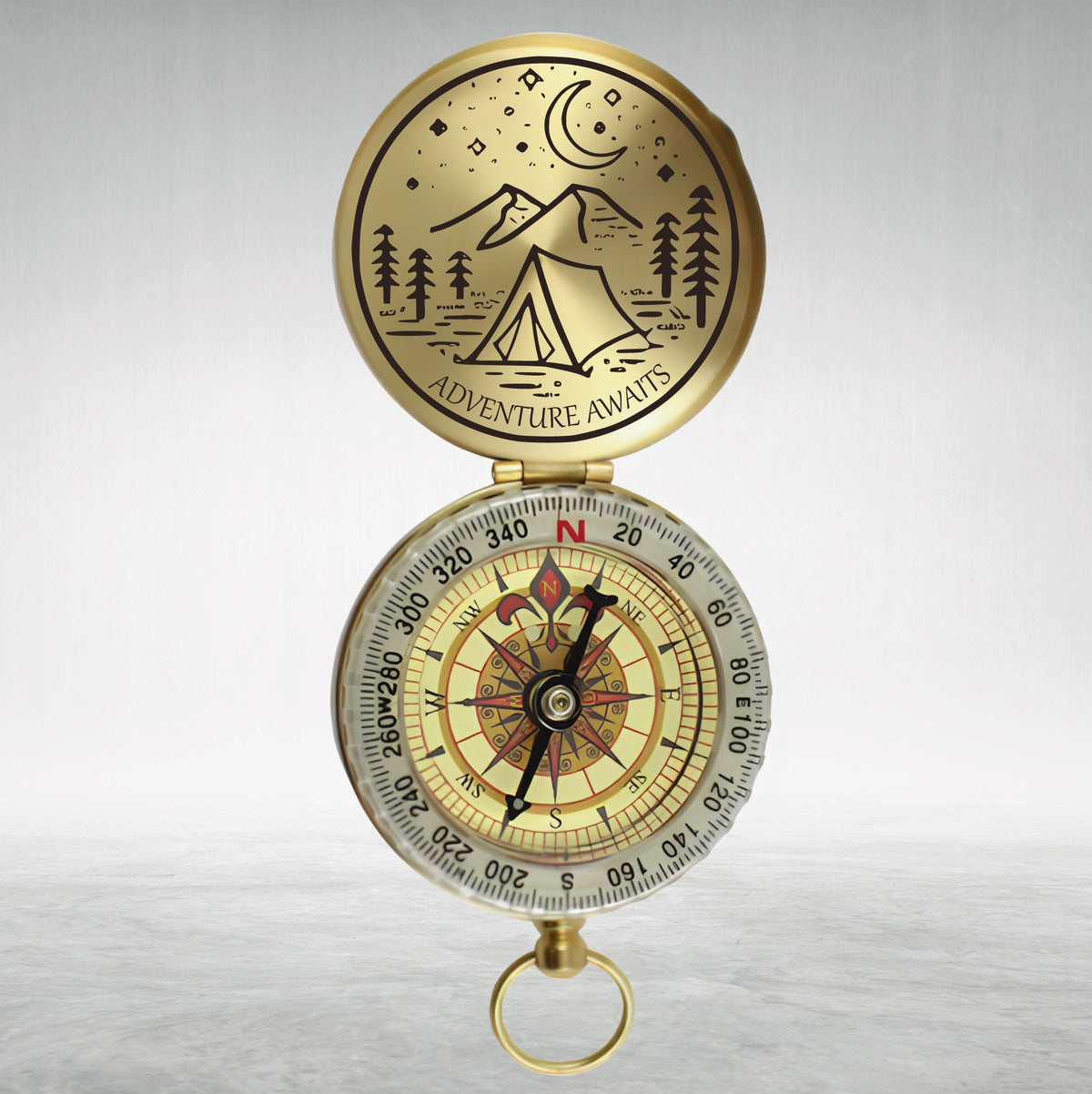 May include: A gold compass with a black needle and a white face. The compass has a lid with a black and white illustration of a tent, mountains, and a crescent moon. The lid has the text "Adventure Awaits".