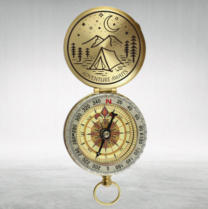 May include: A gold compass with a black needle and a white face. The compass has a lid with a black and white illustration of a tent, mountains, and a crescent moon. The lid has the text "Adventure Awaits".