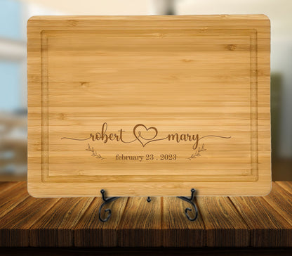 May include: A light brown bamboo cutting board with a personalized inscription. The inscription reads "robert & mary" with a heart symbol and the date "february 23, 2023".