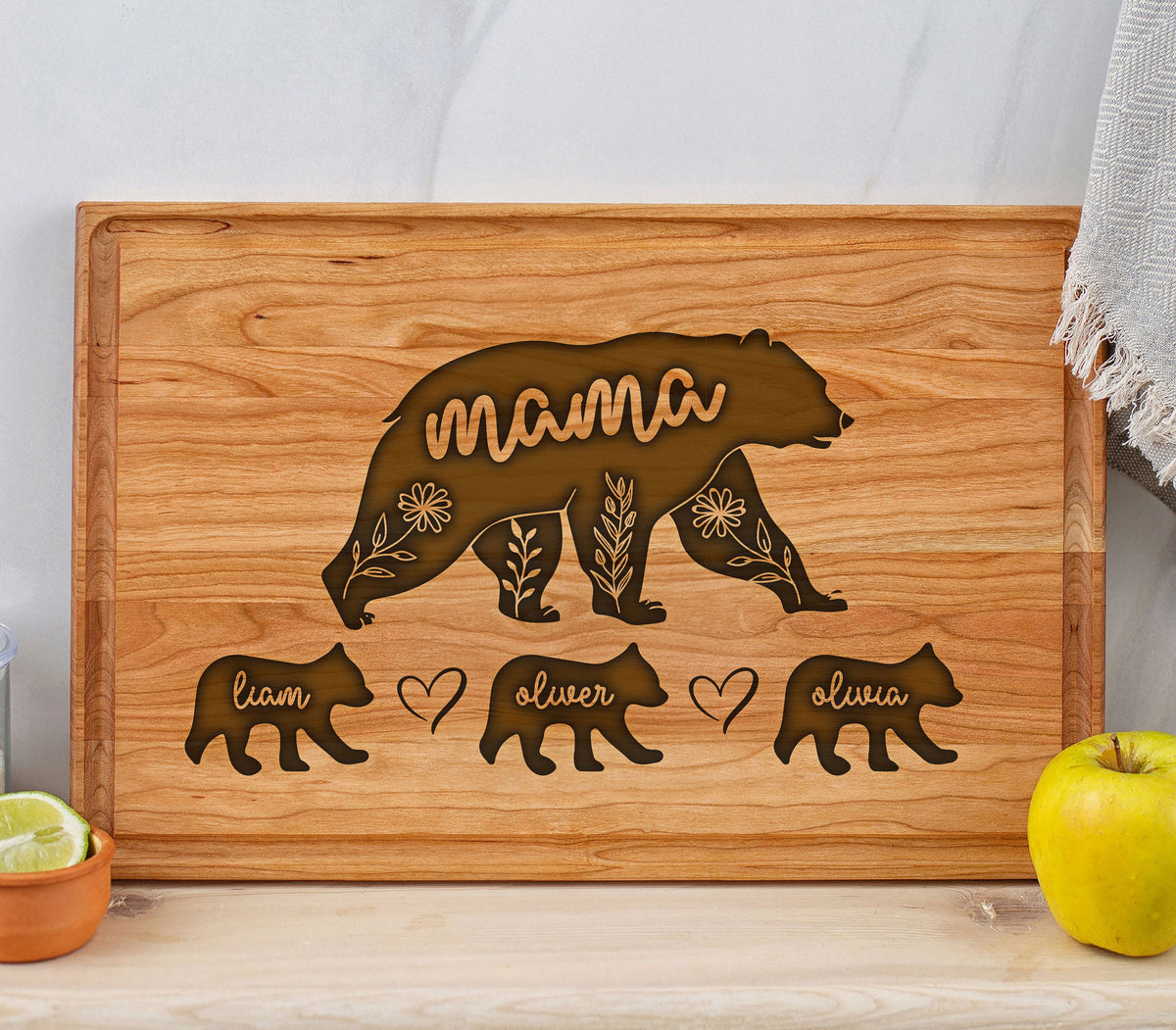 May include: A wooden cutting board with a carved bear design. The word "mama" is carved into the large bear, with three smaller bears below, each with a name. Floral and leaf details are carved into the mama bear.