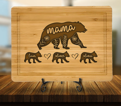 May include: A bamboo cutting board with a carved bear design. The word "mama" is carved into the bear's body, with floral accents. Below are three smaller bears with the names "Liam", "Oliver", and "Olivia". A heart symbol is between "Liam" and "Oliver".