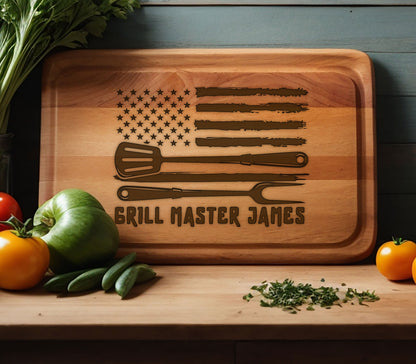 May include: A wooden cutting board with an American flag design and the text "Grill Master James". The flag is brown and the text is in a brown font. There are three grilling tools on the board, a spatula, a knife, and a fork.