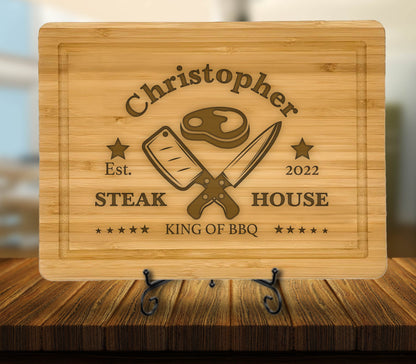 May include: Personalized bamboo cutting board with the text "Christopher Steak House Est. 2022 King of BBQ" and a graphic of crossed butcher knives and a steak.