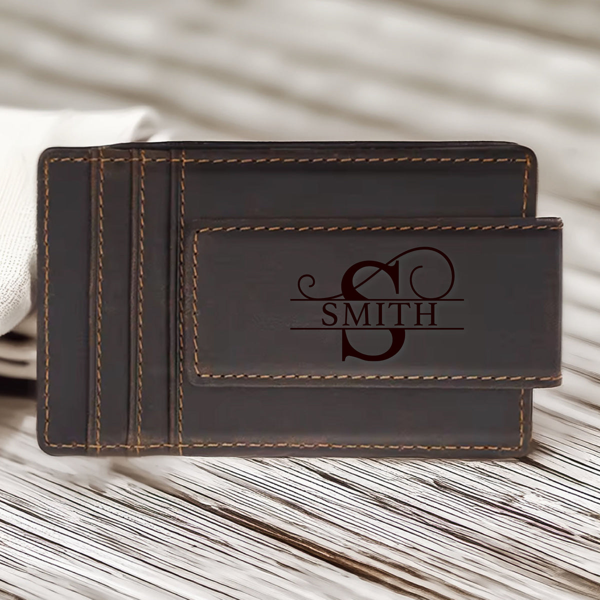 May include: A dark brown leather money clip wallet with a magnetic closure. The wallet features card slots and is personalized with the name "SMITH" and the letter "S". The stitching is a lighter brown color.