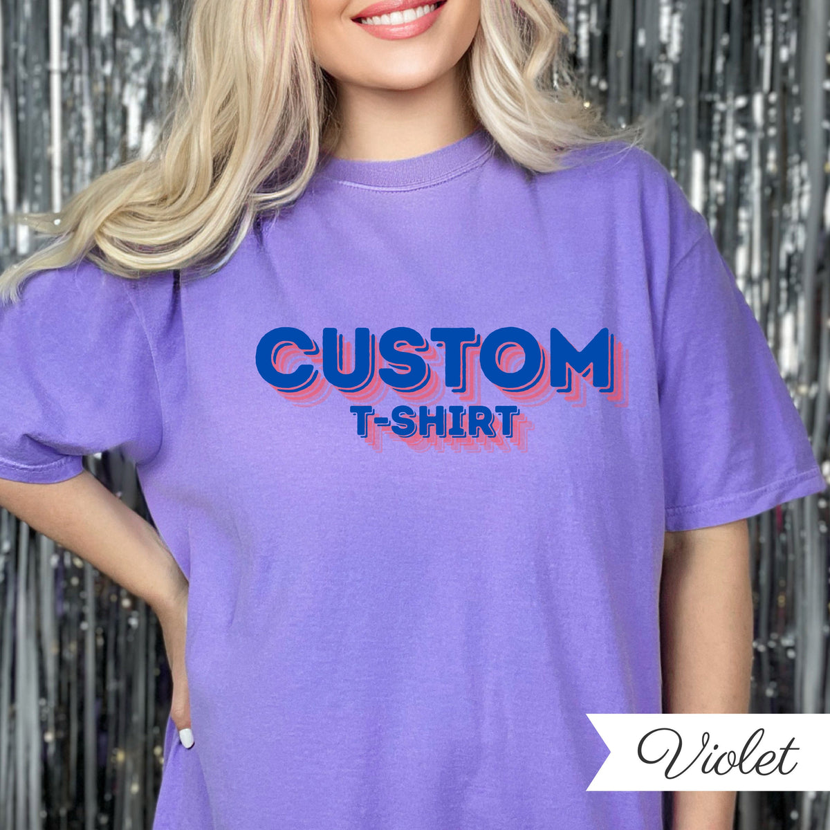 May include: A violet-colored t-shirt with the word "CUSTOM" in blue, outlined in pink, above the words "T-SHIRT". The shirt is a solid color and has a relaxed fit. The word "Violet" is written in cursive in the bottom right corner.