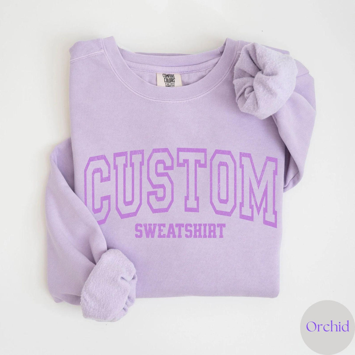 May include: A light purple sweatshirt with the word "CUSTOM" above "SWEATSHIRT" in a matching purple, collegiate-style font. The sleeves are folded, and the sweatshirt is on a white background. The color is labeled "Orchid".