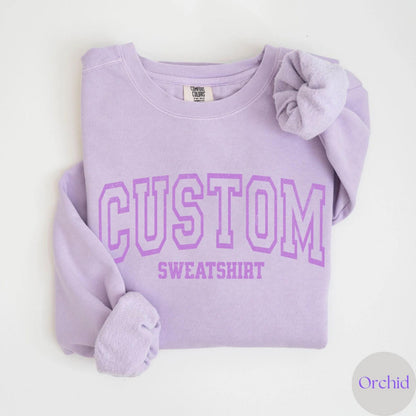 May include: A light purple sweatshirt with the word "CUSTOM" printed in a collegiate font above "SWEATSHIRT". The sweatshirt is a solid color with a crew neck and long sleeves. The color is labeled "Orchid".