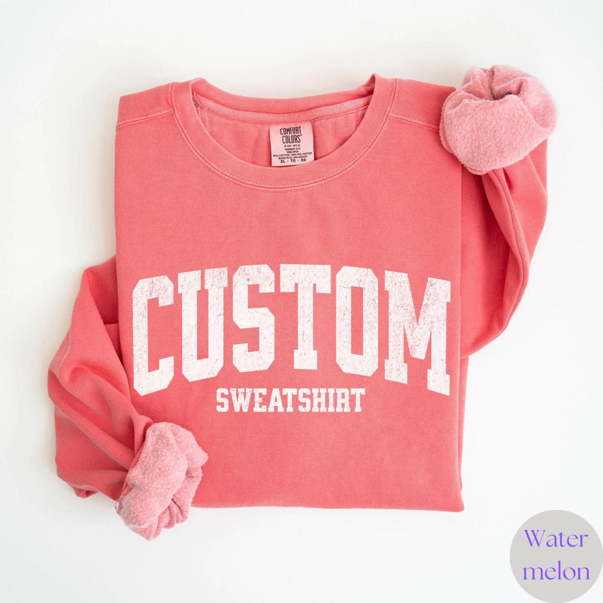May include: A coral-colored sweatshirt with the word "CUSTOM" in large, white, block letters above "SWEATSHIRT". The sleeves are folded, and a matching scrunchie is on the right sleeve. The tag reads "COMFORT COLORS".