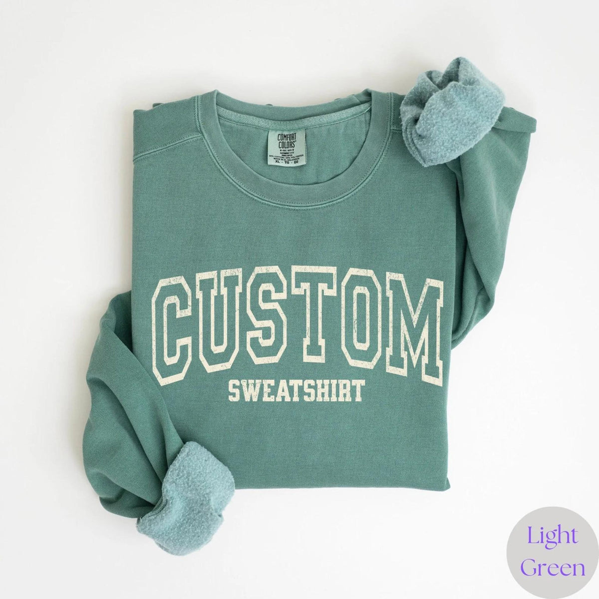 May include: A light green sweatshirt with the word "CUSTOM SWEATSHIRT" printed in white block letters. The sweatshirt is folded, with the sleeves and collar visible. The text "Light Green" is in a small circle in the bottom right corner.
