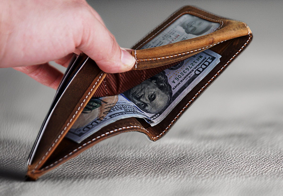 May include: A brown leather wallet with a stitched edge is open, revealing a folded $100 bill.