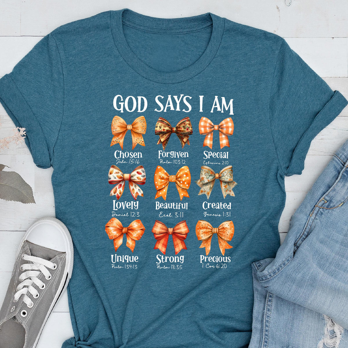 May include: A teal short-sleeved t-shirt featuring a graphic print of nine decorative bows in orange and brown tones. Each bow is paired with a word and a Bible verse, including text such as God Says I Am, Chosen, Forgiven, Special, Lovely, Beautiful, Created, Unique, Strong, and Precious.