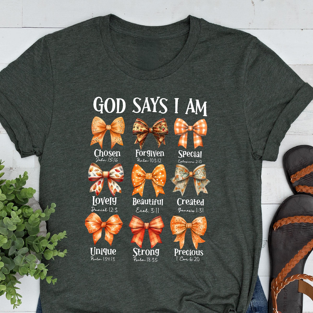 May include: A dark charcoal gray short-sleeve t-shirt featuring the text "GOD SAYS I AM" above nine distinct orange and brown patterned bows. Each bow is paired with a descriptive word and a Bible verse reference printed in white lettering.
