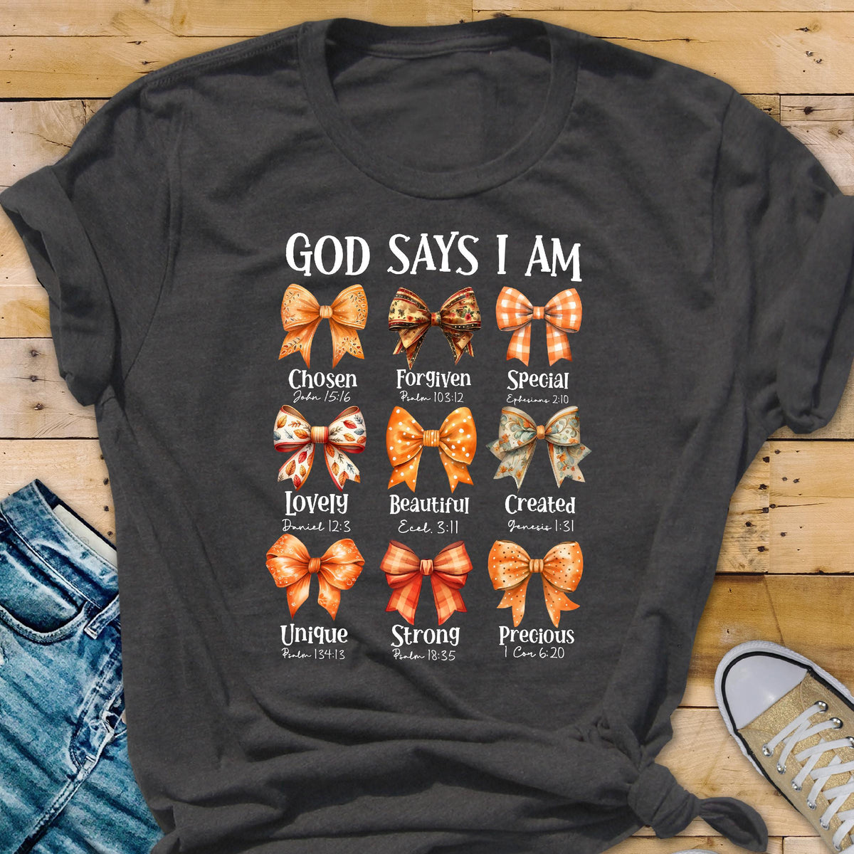May include: A dark gray t-shirt featuring the text GOD SAYS I AM above nine illustrated bows in various orange and brown patterns. Each bow is labeled with a descriptive word and a corresponding Bible verse reference printed in white text.