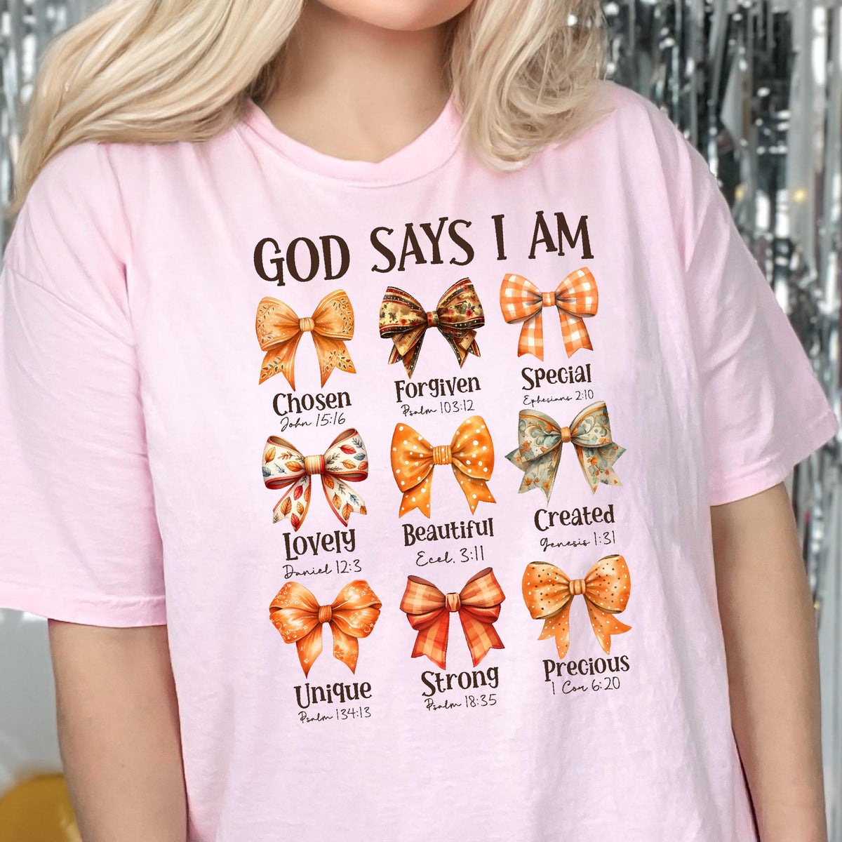 May include: A light pink t-shirt featuring the text GOD SAYS I AM above nine distinct orange and brown patterned bows. Each bow is paired with a descriptive word and a Bible verse reference, including Chosen, Forgiven, Special, Lovely, Beautiful, Created, Unique, Strong, and Precious.