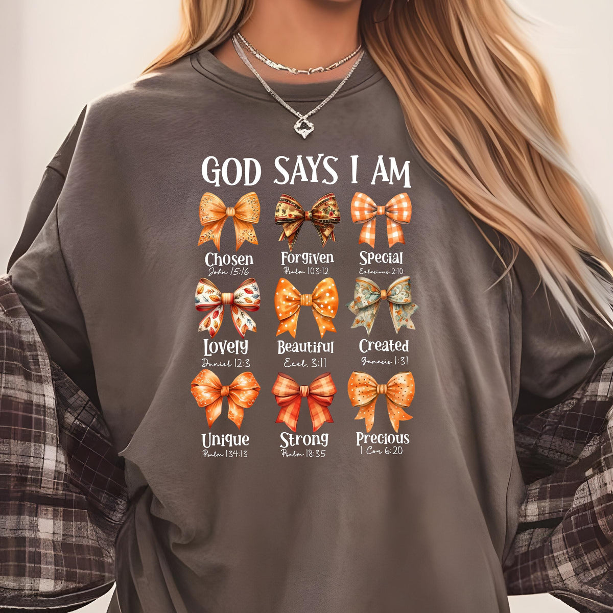 May include: A charcoal gray crewneck sweatshirt featuring the text GOD SAYS I AM above nine illustrated bows in shades of orange, brown, and white. Each bow is paired with a descriptive word and a Bible verse reference printed in white text.
