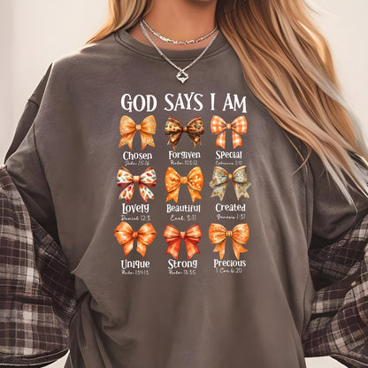 May include: A charcoal gray crewneck sweatshirt featuring the text GOD SAYS I AM above nine illustrated bows in shades of orange, brown, and white. Each bow is paired with a descriptive word and a Bible verse reference printed in white text.