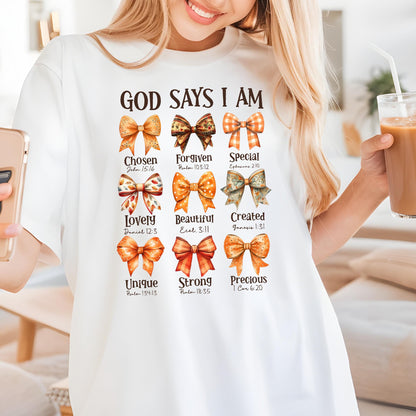 May include: A white t-shirt featuring the text GOD SAYS I AM above nine colorful bows in various orange and brown patterns. Each bow is paired with a descriptive word and a Bible verse reference, including Chosen, Forgiven, Special, Lovely, Beautiful, Created, Unique, Strong, and Precious.