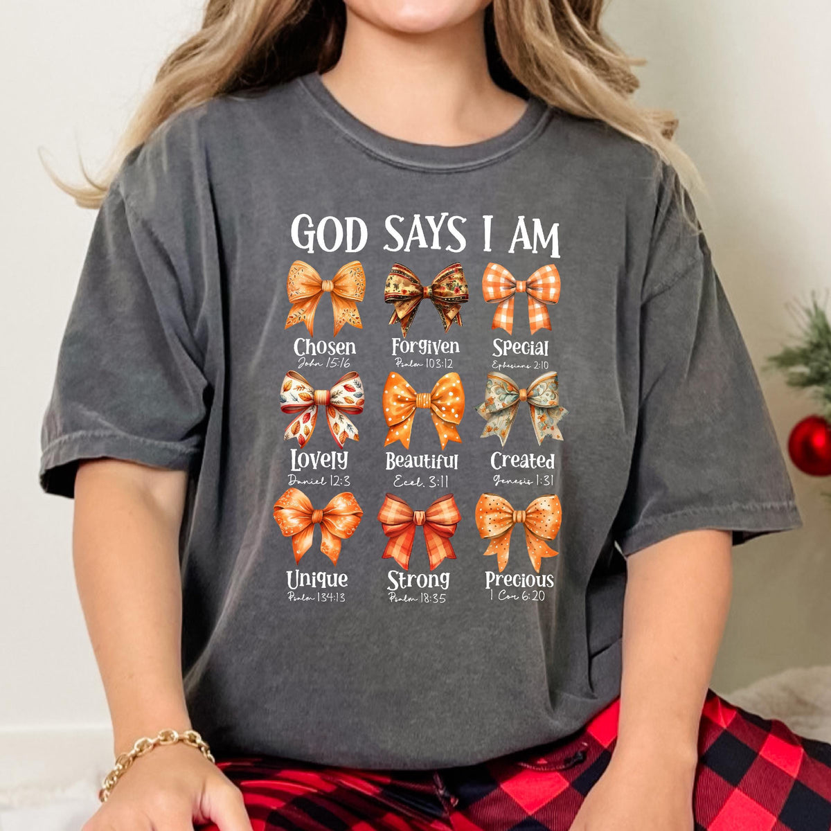 May include: A charcoal gray short-sleeve t-shirt featuring the text "GOD SAYS I AM" above nine illustrated bows in shades of orange and brown. Each bow is paired with a descriptive word and a Bible verse reference, including Chosen, Forgiven, Special, Lovely, Beautiful, Created, Unique, Strong, and Precious.