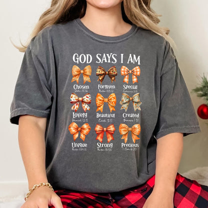 May include: A charcoal gray short-sleeve t-shirt featuring the text "GOD SAYS I AM" above nine illustrated bows in shades of orange and brown. Each bow is paired with a descriptive word and a Bible verse reference, including Chosen, Forgiven, Special, Lovely, Beautiful, Created, Unique, Strong, and Precious.