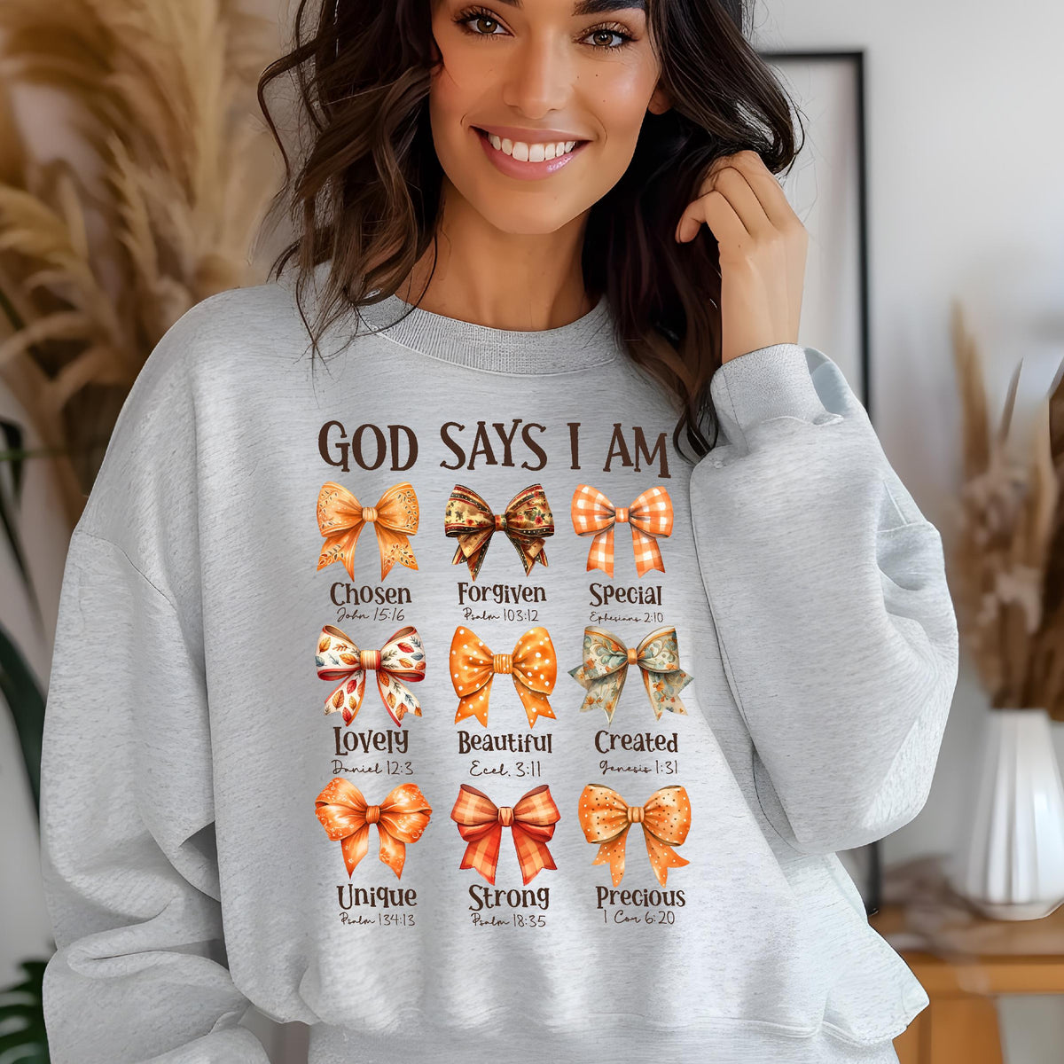 May include: A light gray crewneck sweatshirt featuring the text GOD SAYS I AM at the top, followed by nine distinct illustrated bows in shades of orange, brown, and cream, each paired with a descriptive word and a corresponding Bible verse reference.