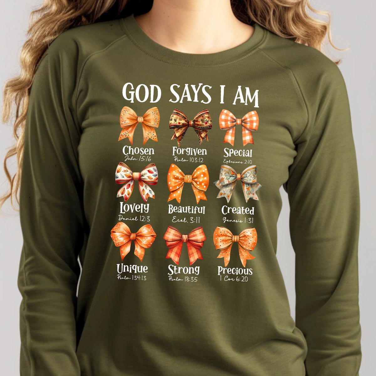 May include: An olive green long-sleeve sweatshirt featuring the text GOD SAYS I AM above nine illustrated bows in various orange and brown patterns. Each bow is labeled with a word and a Bible verse reference, including Chosen, Forgiven, Special, Lovely, Beautiful, Created, Unique, Strong, and Precious.