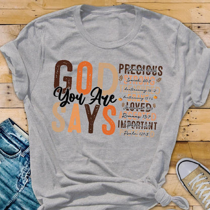 May include: A heather gray t-shirt with the words "GOD SAYS You Are PRECIOUS, LOVED, IMPORTANT" in brown and orange lettering. Bible verses are listed below the words. The shirt is folded on a wooden surface.