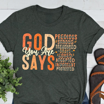 May include: Dark gray t-shirt with the words "GOD You Are SAYS" in orange and cream lettering. The shirt also features a list of words like "Precious" and "Strong" in a smaller font.