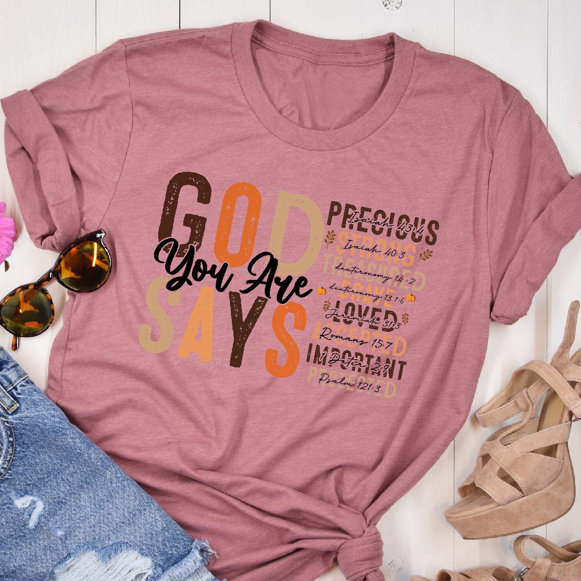 May include: A dusty rose t-shirt with the words "GOD SAYS You Are" in a mix of brown, orange, and beige fonts. The shirt also lists "PRECIOUS, LOVED, IMPORTANT" with Bible verses.