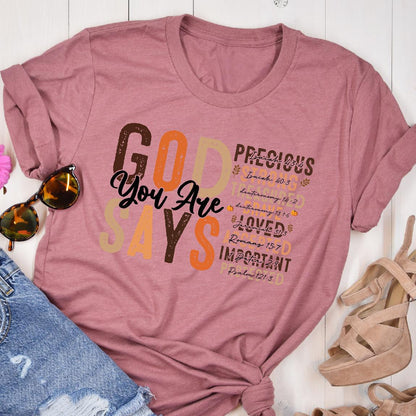 May include: A dusty rose t-shirt with the words "GOD SAYS You Are" in a mix of brown, orange, and beige fonts. The shirt also lists "PRECIOUS, LOVED, IMPORTANT" with Bible verses.