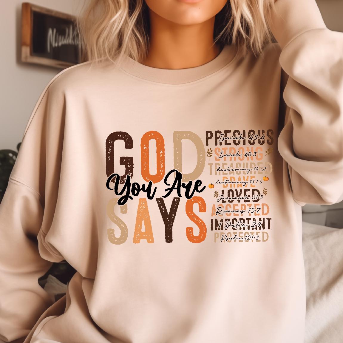 May include: A beige sweatshirt with a graphic print that says "GOD YOU ARE SAYS" in brown, orange, and white. The graphic also includes the words "PRECIOUS", "STRONG", "TREASURED", "BRAVE", "LOVED", "ACCEPTED", "IMPORTANT", and "PROTECTED" with corresponding bible verses.