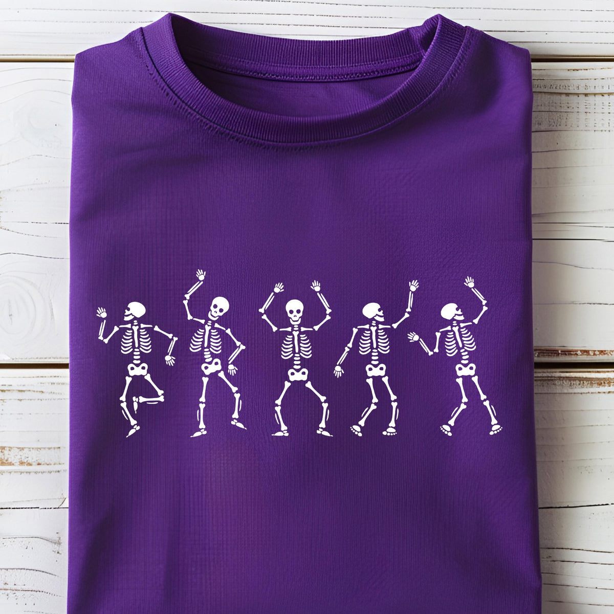 May include: A purple t-shirt featuring a white graphic of five dancing skeletons. The skeletons are in various poses with their arms raised. The shirt is folded neatly on a white wooden surface.