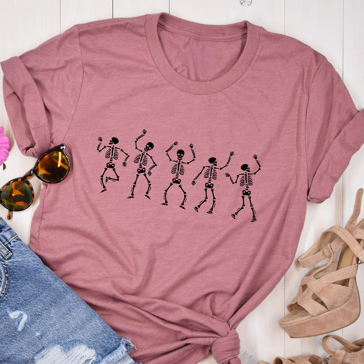 May include: A dusty rose-colored t-shirt featuring a black graphic of five dancing skeletons. The skeletons are in various poses with their arms raised. The shirt is made of a soft material and has a crew neck.
