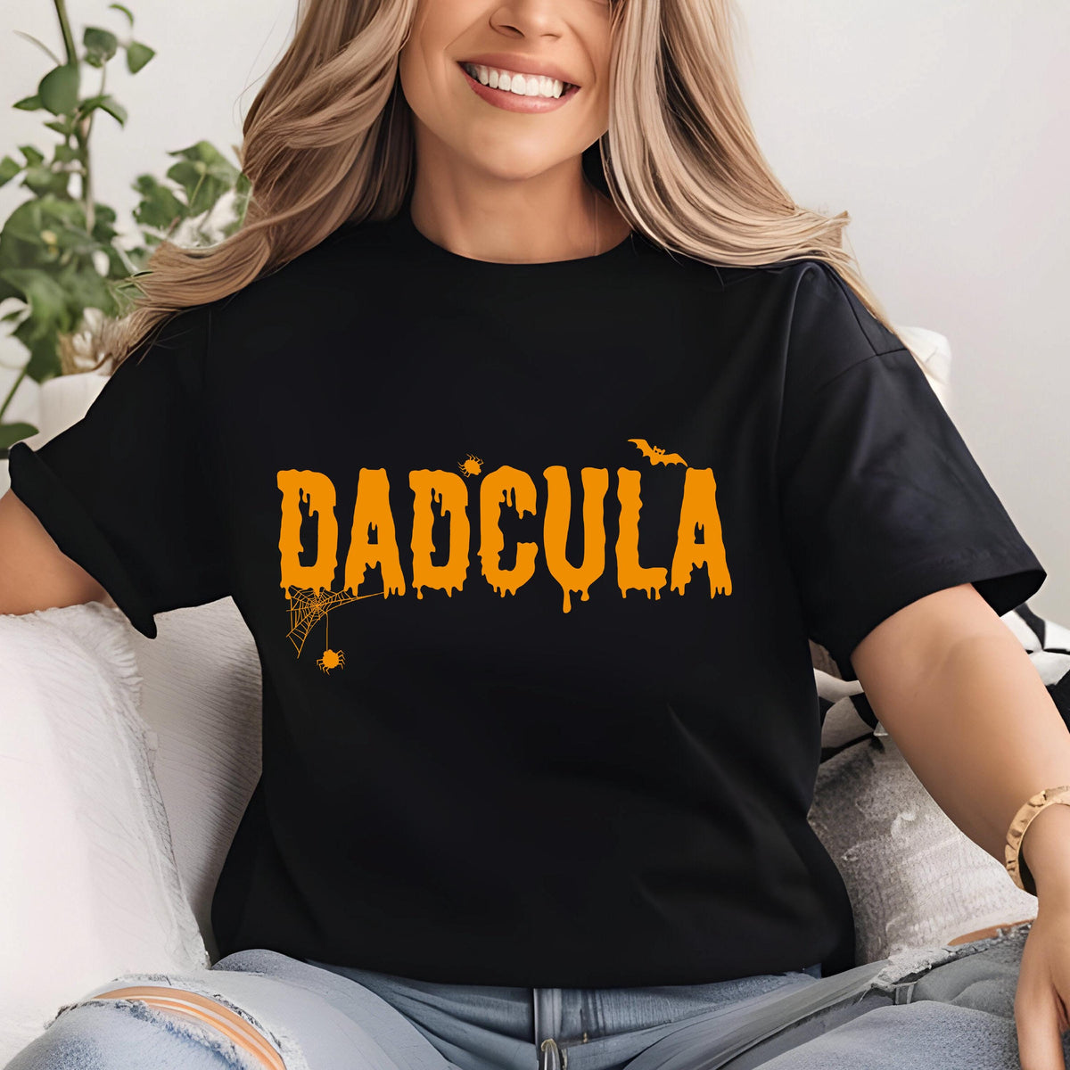 May include: A black short-sleeved t-shirt featuring the orange text DADCULA in a dripping font. The design includes a small spider web with a spider hanging from the letter D, a small bat above the letter L, and two small spider icons.