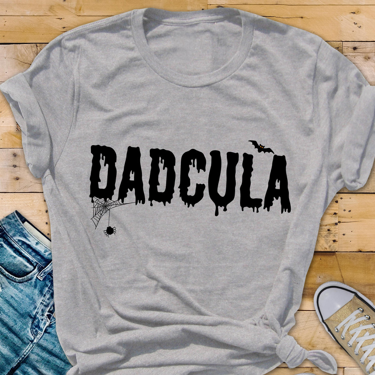 May include: A light gray short-sleeved t-shirt featuring the black text DADCULA in a dripping font. The design includes a small black spider hanging from a web attached to the letter D and a small black bat silhouette above the letter A.