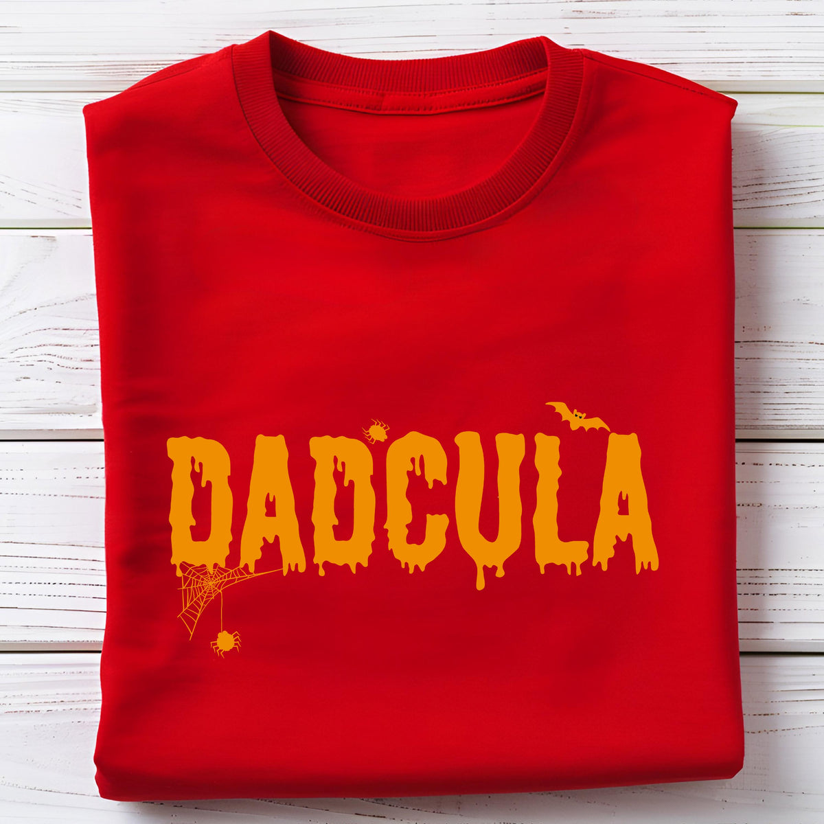 May include: A bright red crew neck t-shirt featuring the orange text DADCULA in a dripping, horror-inspired font. The design includes a small orange spider hanging from a web on the left side, a tiny spider above the letters, and a bat.