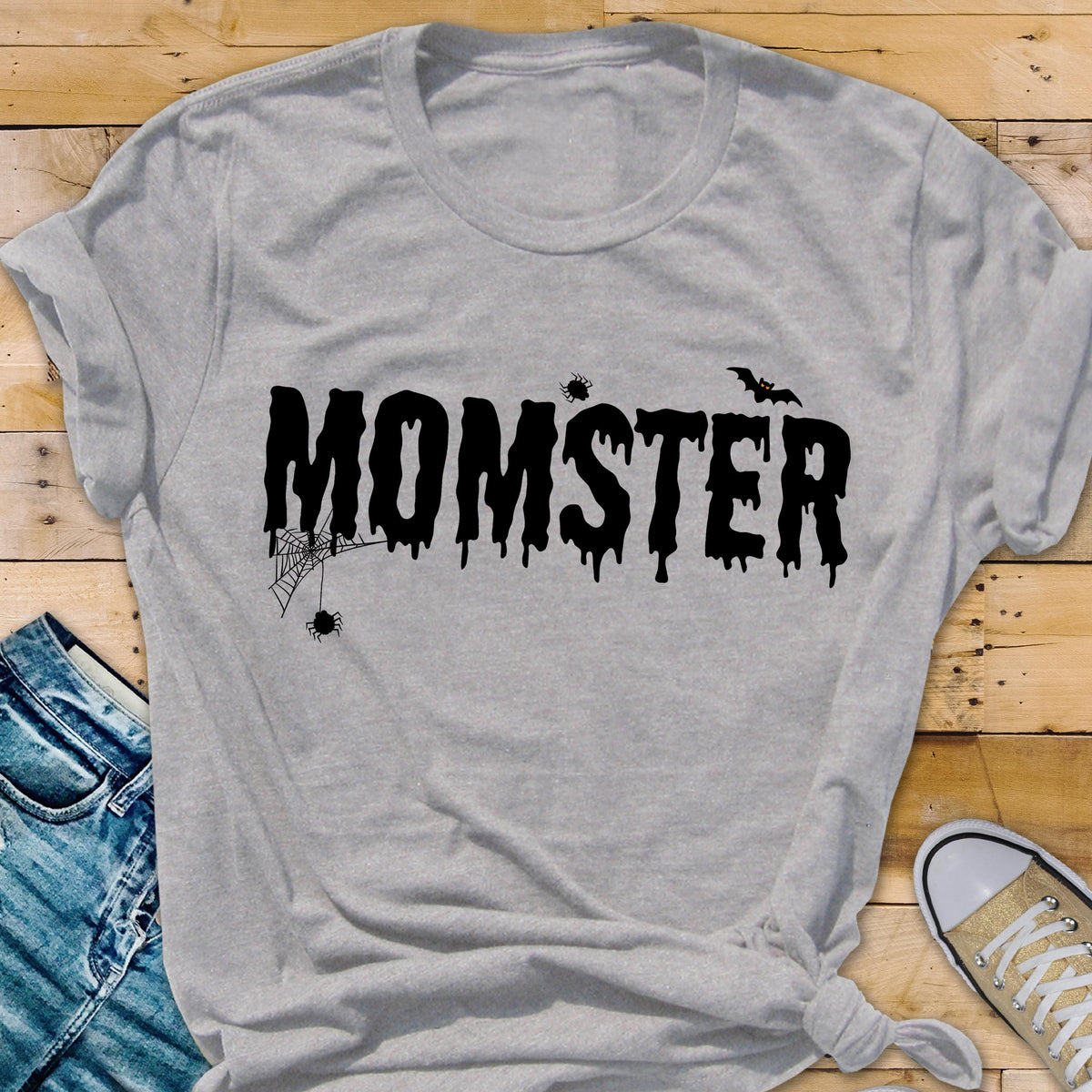 May include: A light gray t-shirt with the word "MOMSTER" in black, dripping font. The design includes a spider web, spiders, and a bat, creating a Halloween theme. The shirt is knotted at the bottom.