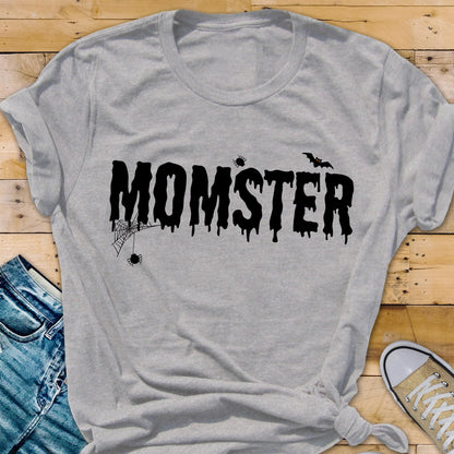 May include: A light gray t-shirt with the word "MOMSTER" in black, dripping font. The design includes a spider web, spiders, and a bat, creating a Halloween theme. The shirt is knotted at the bottom.