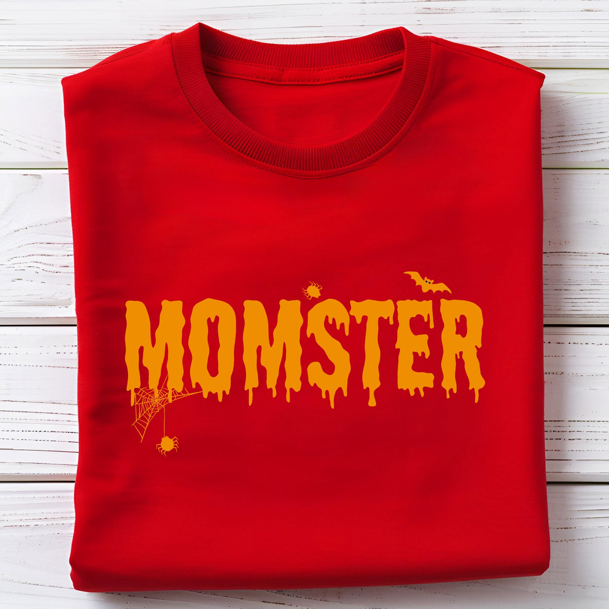 May include: A red sweatshirt with the word "MOMSTER" in orange dripping letters. The design includes a spider web, spiders, and a bat, all in orange. The sweatshirt is folded neatly on a white wooden surface.