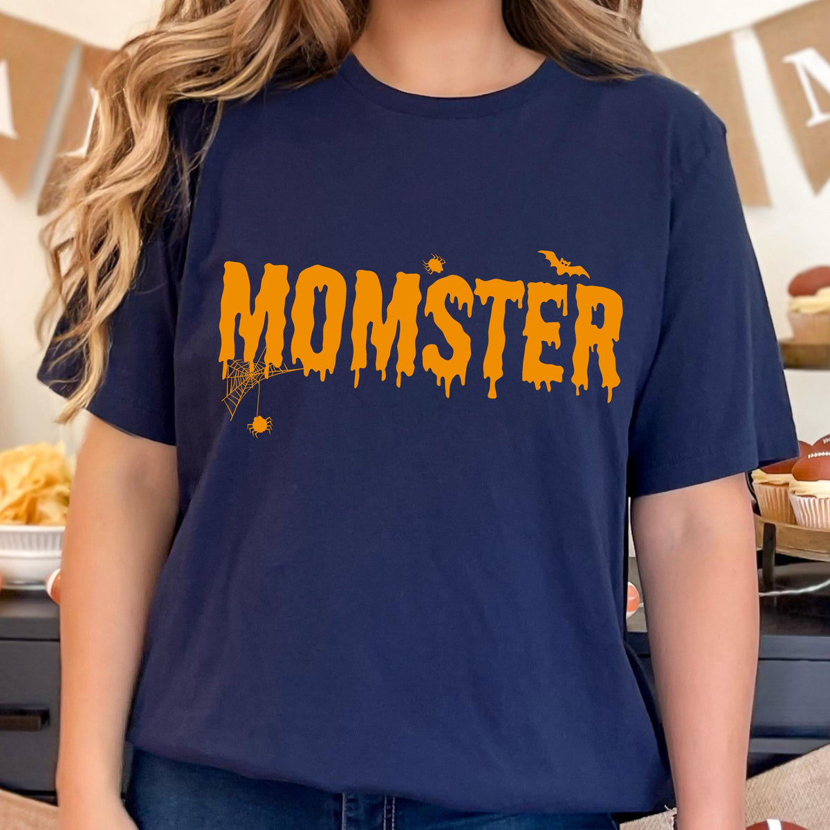 May include: Navy blue t-shirt with the word "MOMSTER" in orange dripping font. A spider web and bat graphic are above and below the text. The shirt is a Halloween-themed design.